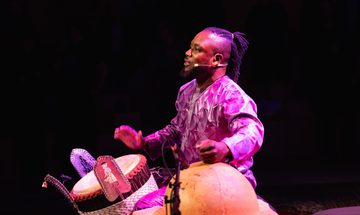 A man seated on stage, illuminated by purple light, playing two percussion instruments