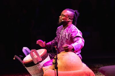 A man seated on stage, illuminated by purple light, playing two percussion instruments