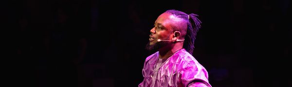 A man seated on stage, illuminated by purple light, playing two percussion instruments