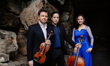 Two men and a woman stand in front of a large rock. The men wear black suits and the woman wears a bright blue dress. They are holding a violin and a cello.