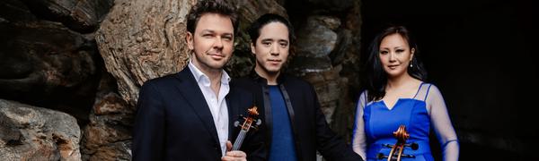 Two men and a woman stand in front of a large rock. The men wear black suits and the woman wears a bright blue dress. They are holding a violin and a cello.