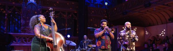 A woman playing the double bass performing on stage with two brass players and a drummer