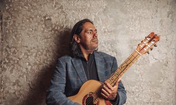 A man stands in front of a stone wall holding a guitar and looking to the side.