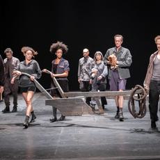 An ensemble of performers walk across a stage holding a wooden prop. They are all wearing different shades of grey.