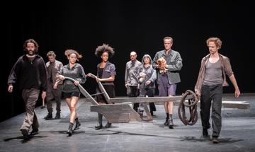 An ensemble of performers walk across a stage holding a wooden prop. They are all wearing different shades of grey.