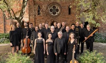 A group of musicians dressed in black standing in front of a brick building. Trees and bushes frame them.