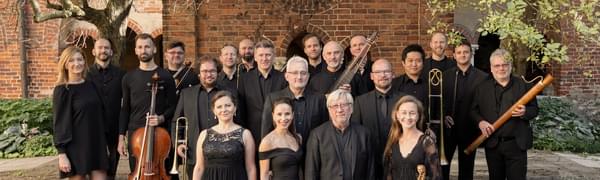 A group of musicians dressed in black standing in front of a brick building. Trees and bushes frame them.
