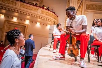 a boy sits on stage holding a violin smiling at his mum