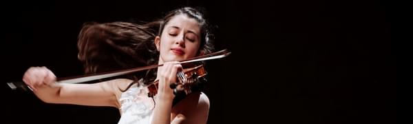 A young woman in a white dress plays the violin, her long brown hair arcs through the air behind her