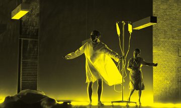 A black and yellow image of an actor wearing a hospital gown and holding an IV drip. A woman stands behind them while another person lays on the floor in front of them.
