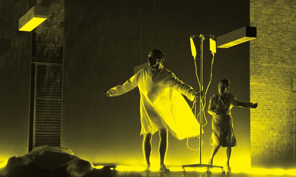 A black and yellow image of an actor wearing a hospital gown and holding an IV drip. A woman stands behind them while another person lays on the floor in front of them.