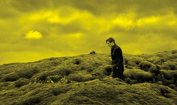 A black and yellow image of Vikingur Olafsson walking through a field with a cloudy sky behind him