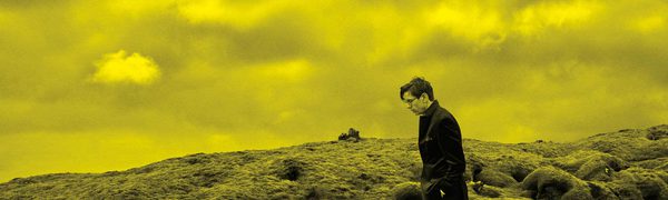 A black and yellow image of Vikingur Olafsson walking through a field with a cloudy sky behind him