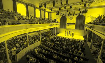 A black and yellow photo of a quartet performing on stage while an audience watches in the Queens Hall