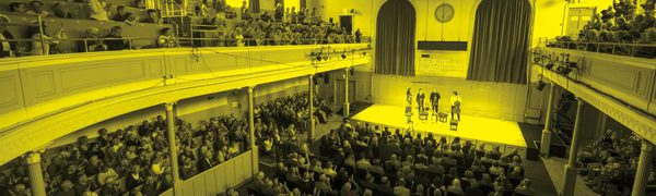 A black and yellow photo of a quartet performing on stage while an audience watches in the Queens Hall