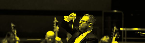 A black and yellow image of a conductor holding a baton and standing in front of the orchestra