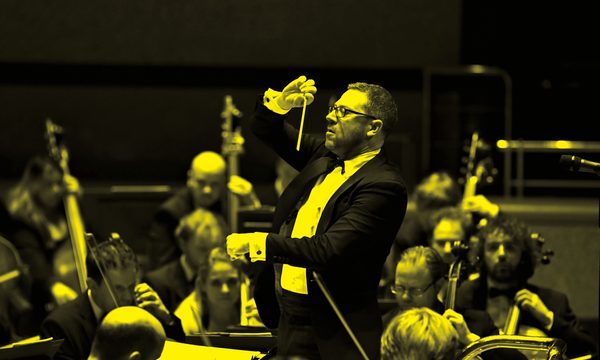 A black and yellow image of a conductor holding a baton and standing in front of the orchestra