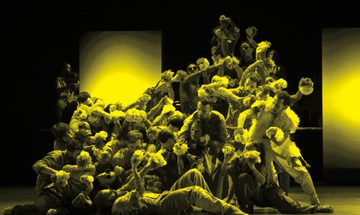 A black and yellow image of an ensemble of dancers moving across the stage
