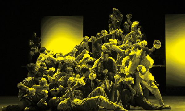 A black and yellow image of an ensemble of dancers moving across the stage