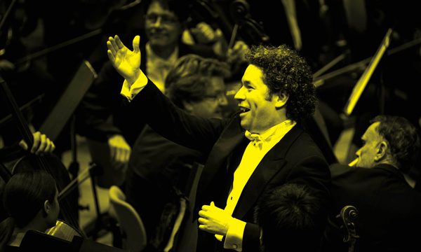 A black and yellow image of a conductor standing in front of an orchestra with his arm in the air while smiling