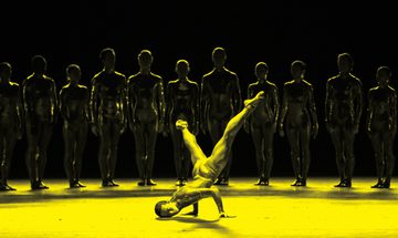 A black and yellow image of a dancer balancing on their hands on stage, while other dancers stand in a line behind them