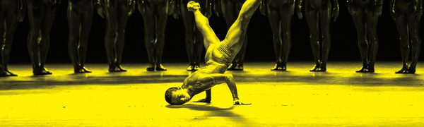 A black and yellow image of a dancer balancing on their hands on stage, while other dancers stand in a line behind them