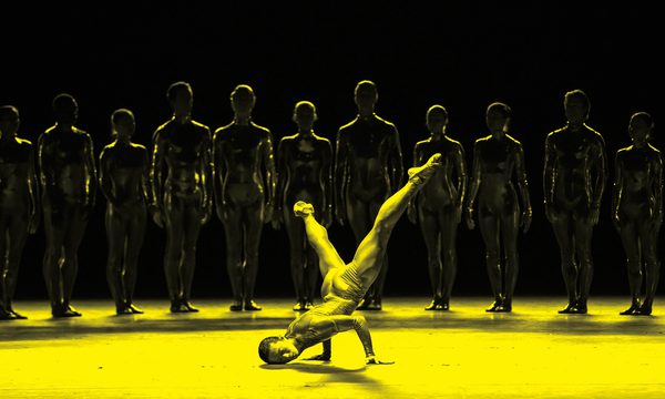 A black and yellow image of a dancer balancing on their hands on stage, while other dancers stand in a line behind them