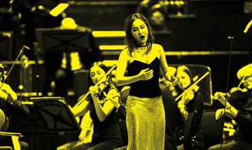 A black and yellow image of an opera singer singing on stage while an orchestra plays behind her