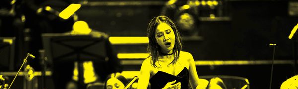 A black and yellow image of an opera singer singing on stage while an orchestra plays behind her