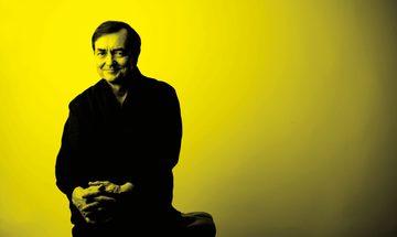 A black and yellow image of Pierre Laurent Aimard sitting on a stool and smiling at the camera