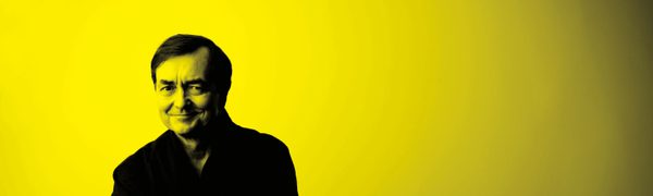 A black and yellow image of Pierre Laurent Aimard sitting on a stool and smiling at the camera