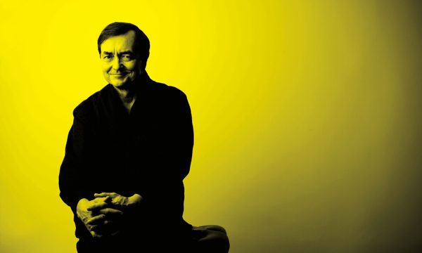 A black and yellow image of Pierre Laurent Aimard sitting on a stool and smiling at the camera