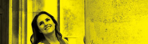 A black and yellow image of Louise Alder leaning against a wall and smiling