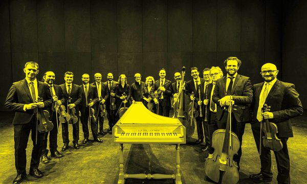 A black and yellow image of the Venice Baroque Orchestra standing in a row holding their instruments