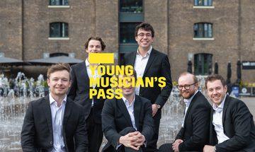 A yellow Young Musician's Pass logo in front of six men wearing suits sit and stand around a fountain