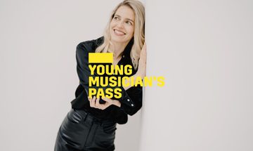 A yellow Young Musician's Pass logo in front of a woman dressed in black leans against a white wall while smiling into the distance
