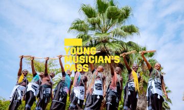 A yellow Young Musician's Pass logo in front of a group of women with vibrantly coloured hair stand in a row holding up drumsticks