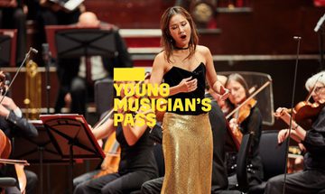 A yellow Young Musician's Pass logo in front of a woman singing on stage wearing a black top and a gold sparkly skirt while an orchestra plays behind her