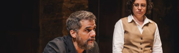 A middle-aged man in a black suit leans forward at a desk, speaking to someone out of frame. A woman in a shirt and vest stands next to him.