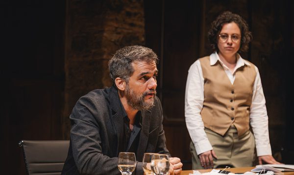 A middle-aged man in a black suit leans forward at a desk, speaking to someone out of frame. A woman in a shirt and vest stands next to him.
