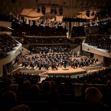 An orchestra performs in a modern concert hall. The audience is sat listening from all sides of the stage.