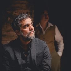 A middle-aged man in a black suit looks off to the side. A woman in a vest and shirt stands in the background behind him