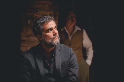 A middle-aged man in a black suit looks off to the side. A woman in a vest and shirt stands in the background behind him