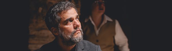 A middle-aged man in a black suit looks off to the side. A woman in a vest and shirt stands in the background behind him