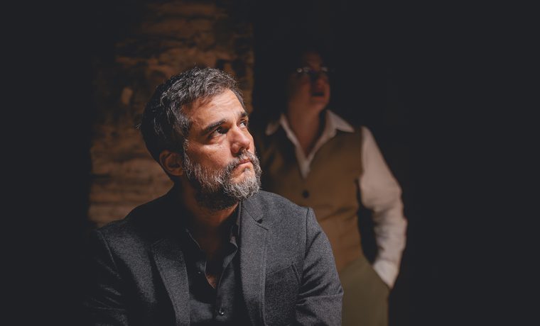 A middle-aged man in a black suit looks off to the side. A woman in a vest and shirt stands in the background behind him