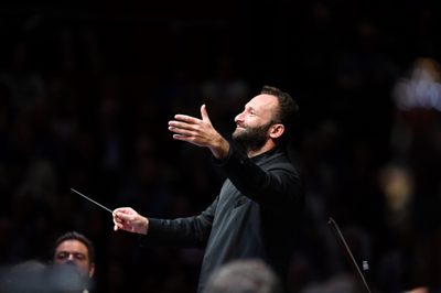A bearded conductor smiles and holds his hand out