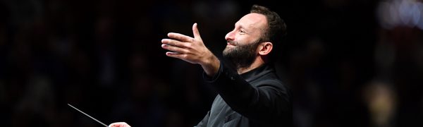 A bearded conductor smiles and holds his hand out