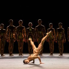 A dancer in a skin tight costume breakdances while a line of other dances watches from the shadows