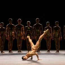 A dancer in a skin tight costume breakdances while a line of other dances watches from the shadows