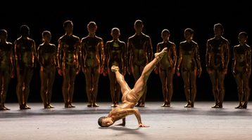 A dancer in a skin tight costume breakdances while a line of other dances watches from the shadows